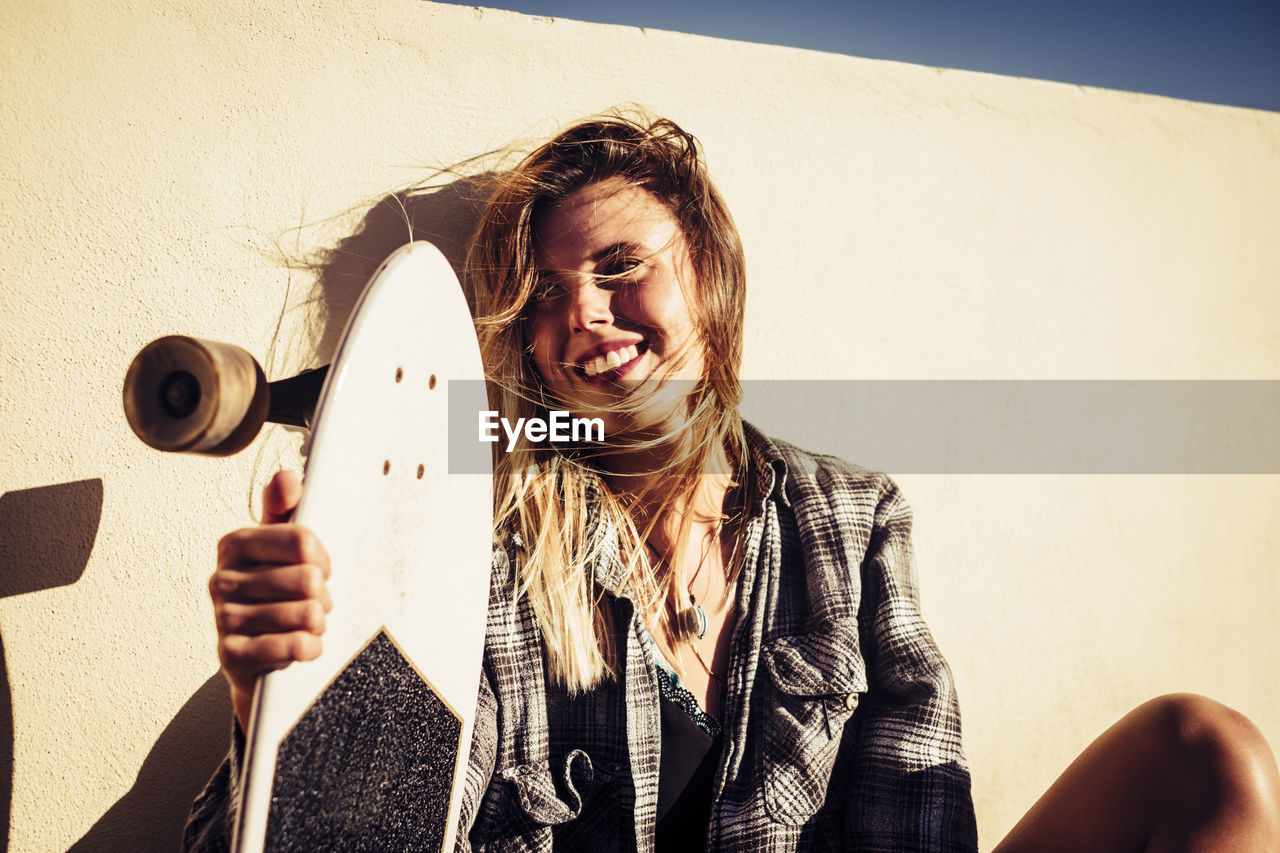 Close-up of young woman holding skateboard by wall