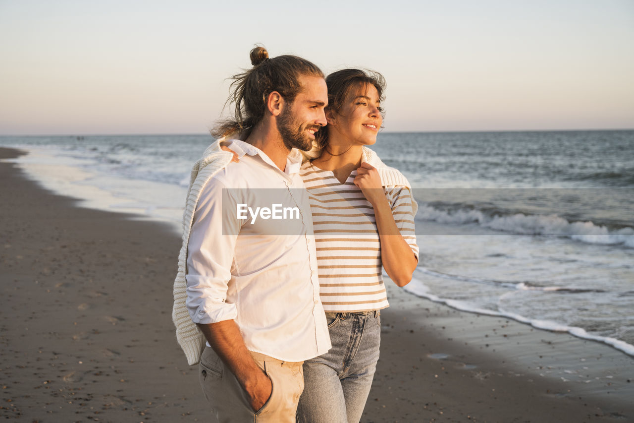 Smiling young couple looking away while walking at beach during vacation