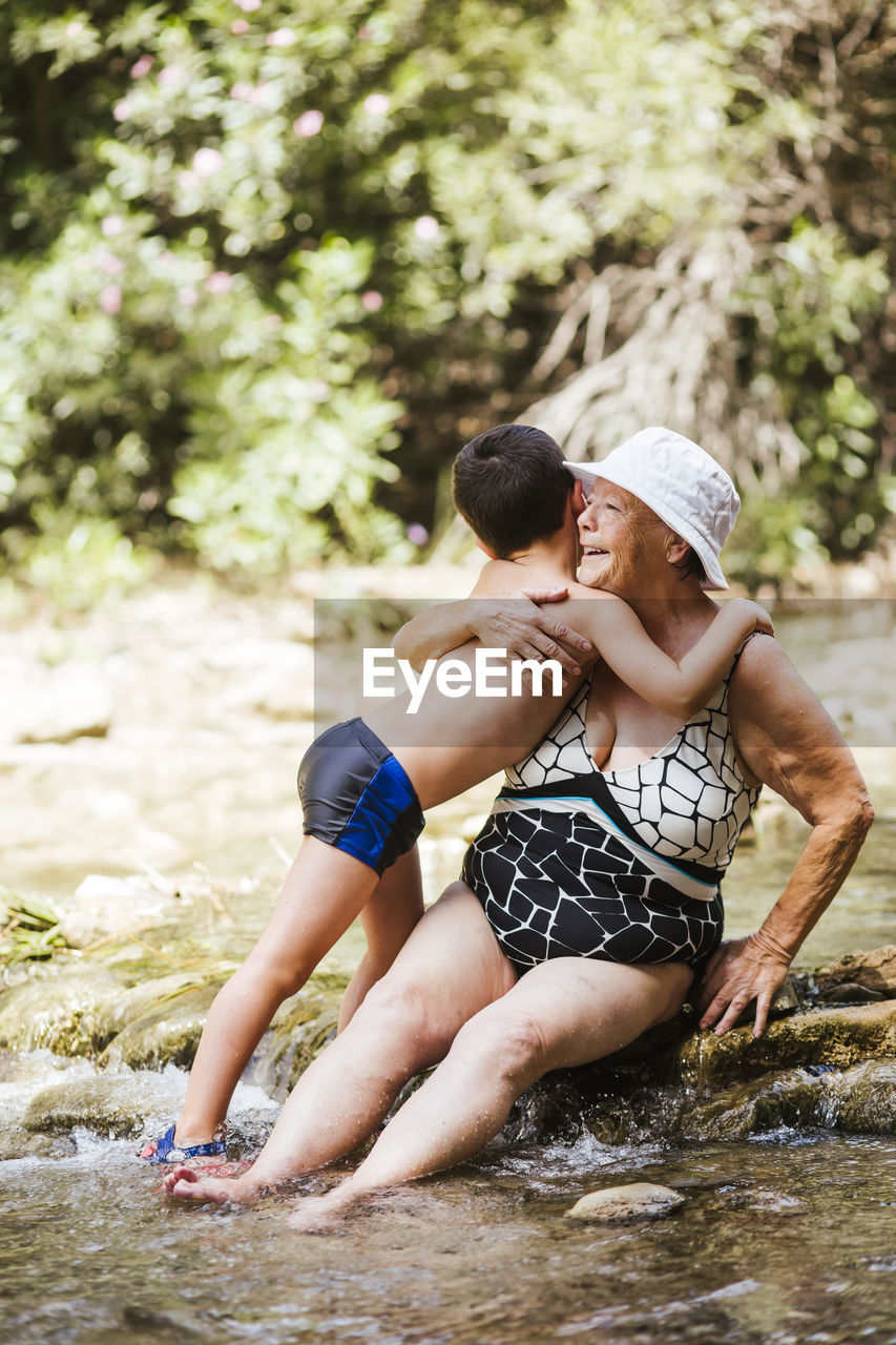 Grandson embracing grandson while sitting on stone in river at forest