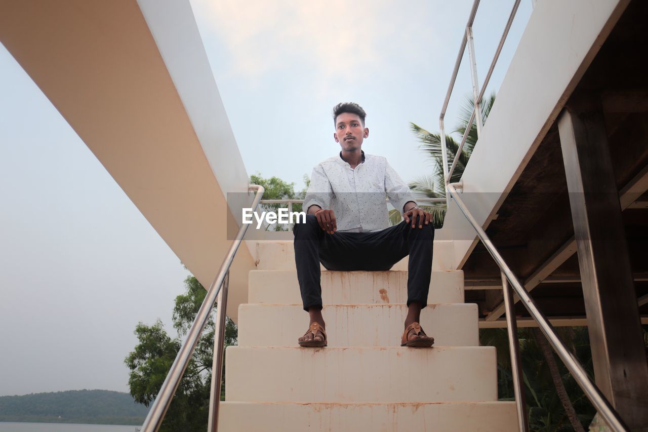 Low angle view portrait of young man sitting on staircase against sky