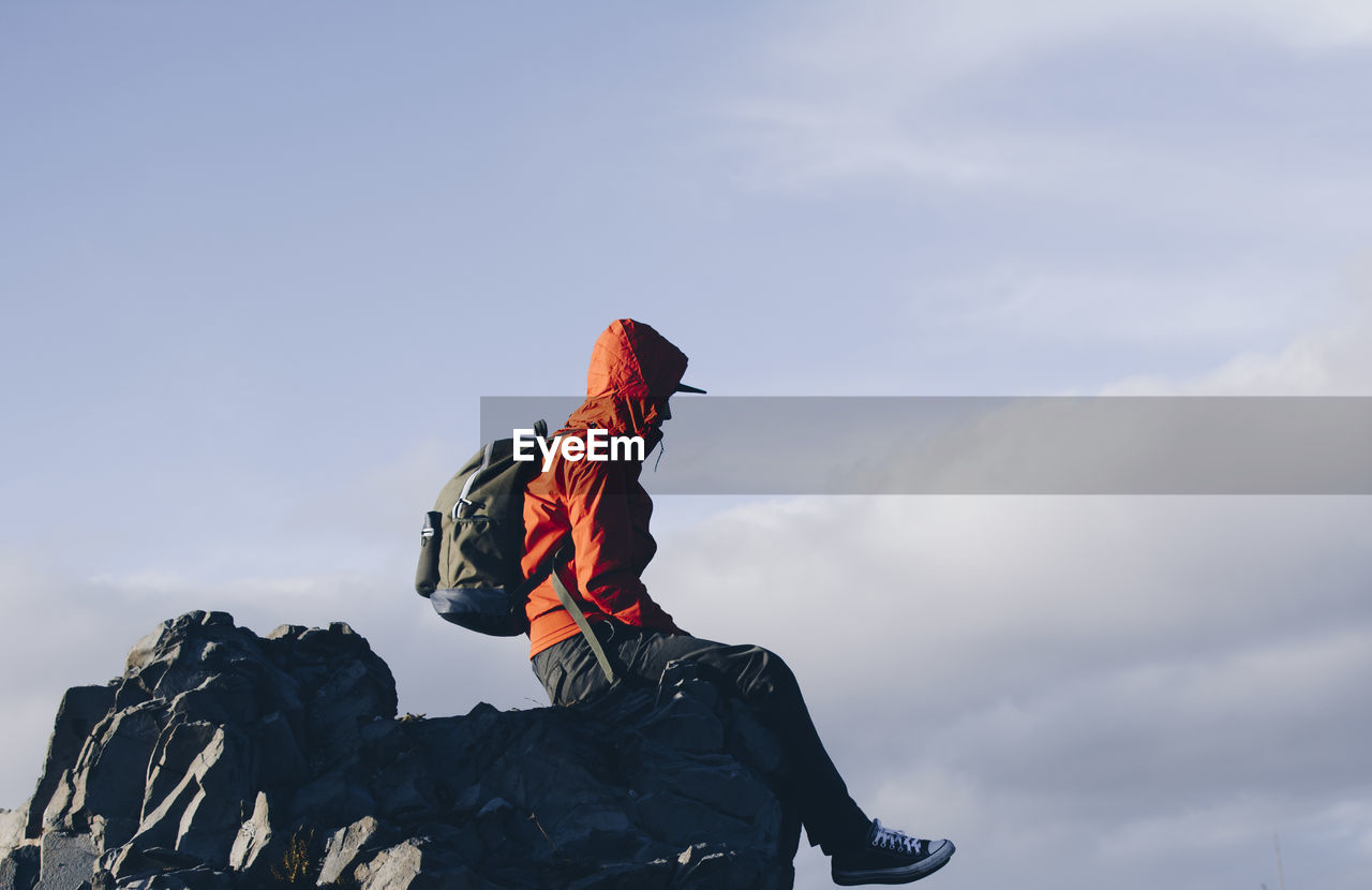 Side view of mid adult man with backpack sitting on rock formation against cloudy sky