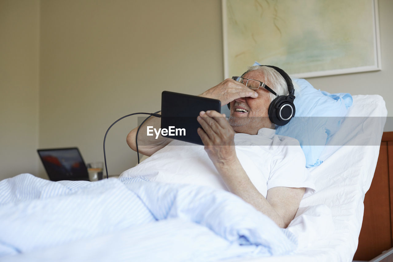 Senior man rubbing eyes while using digital tablet on bed in hospital ward
