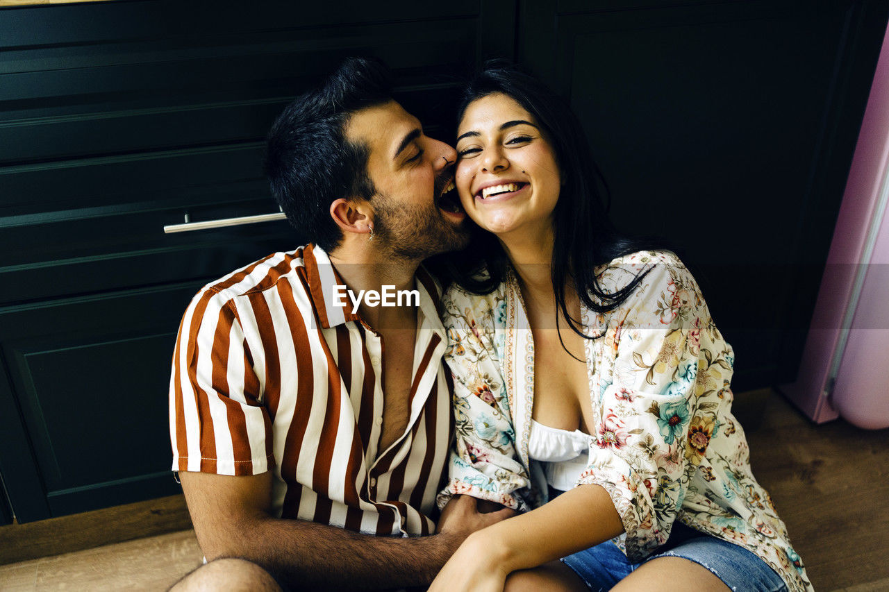 Romantic man biting cheek of happy girlfriend sitting at home