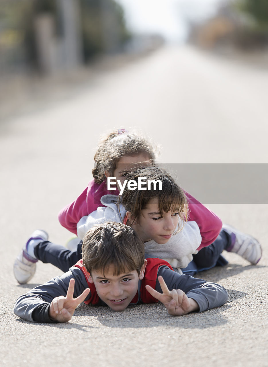 Portrait of boy with sisters showing peace signs while lying down on road