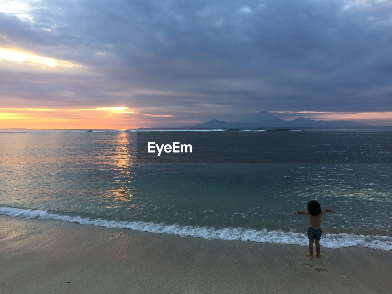 Rear view of shirtless child standing on shore at beach against cloudy sky during sunset