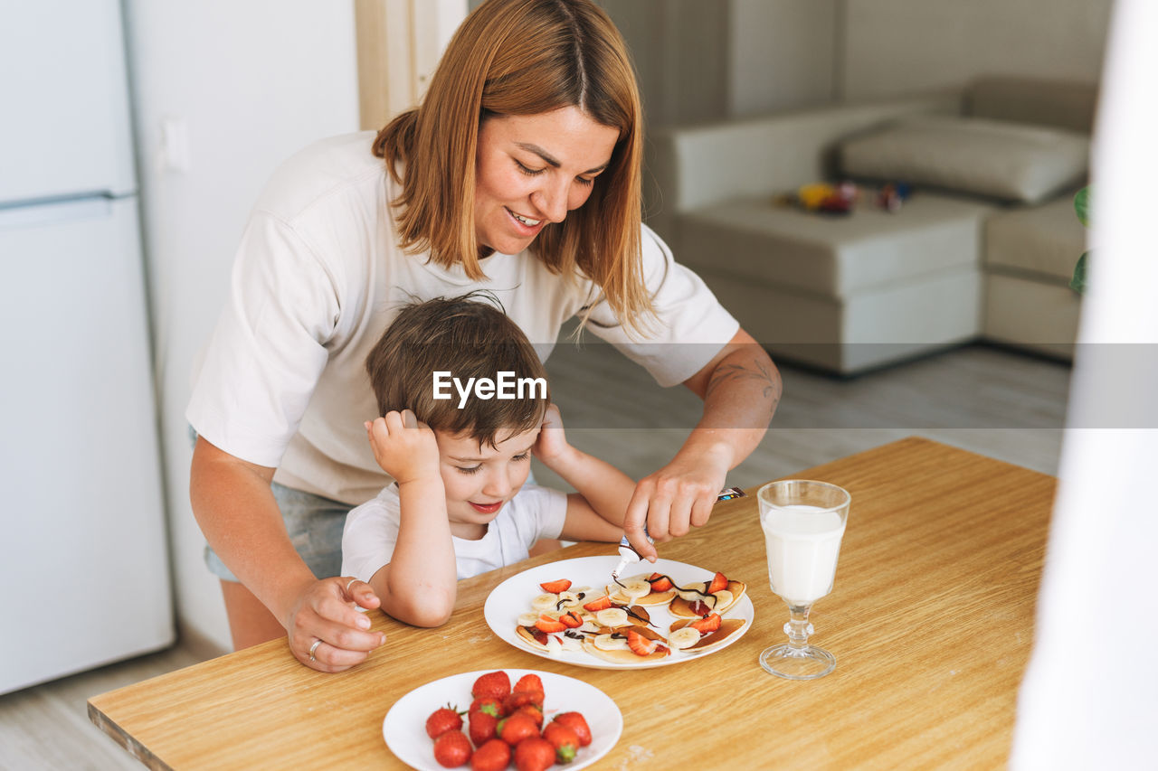 Cute toddler boy with mother having breakfast with puncakes and berries and glass of milk in kitchen 