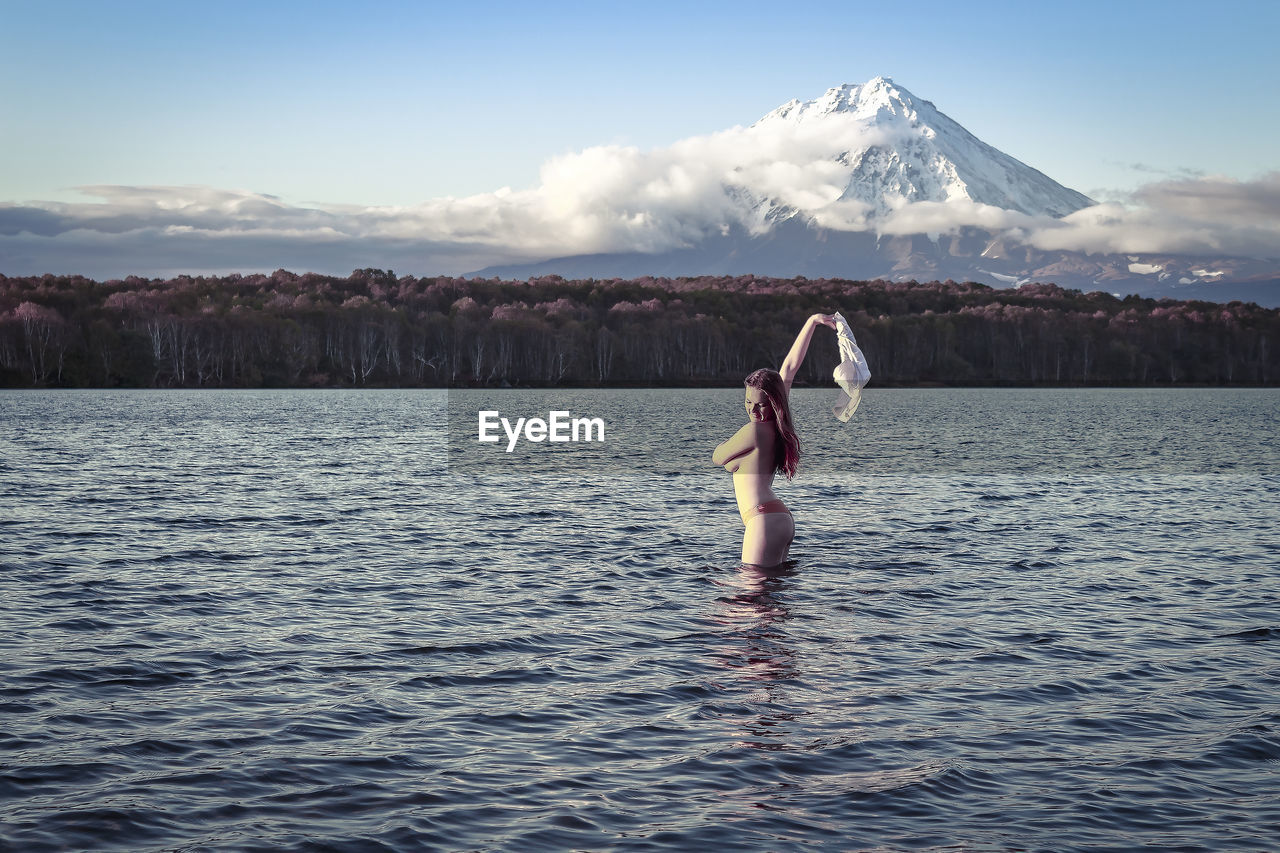 Side view of topless young woman standing in lake against sky during sunset