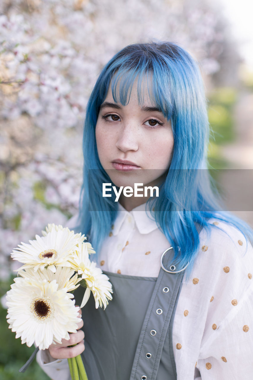 Modern trendy female with blue hair holding bouquet of fresh flowers and looking at camera while standing in blooming spring garden