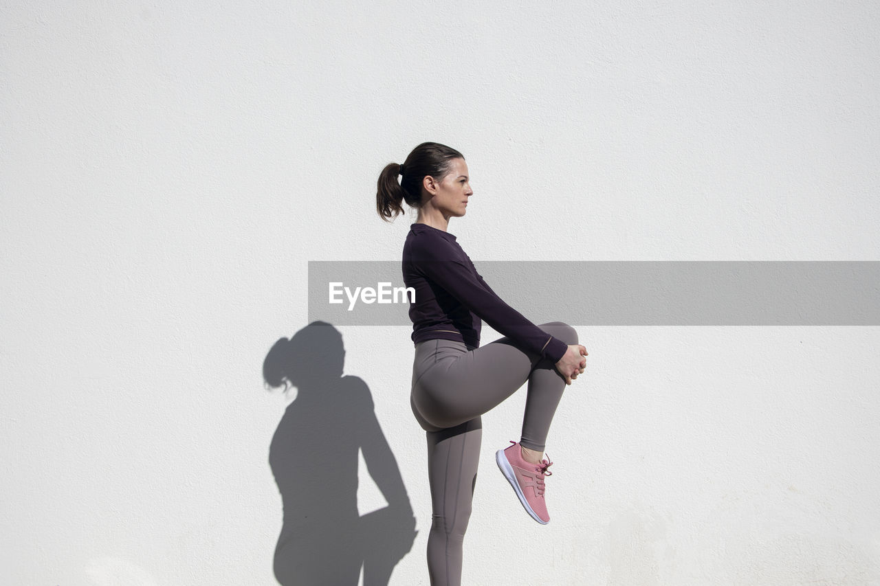 Fit sporty woman doing leg stretching exercises outside against a white wall.