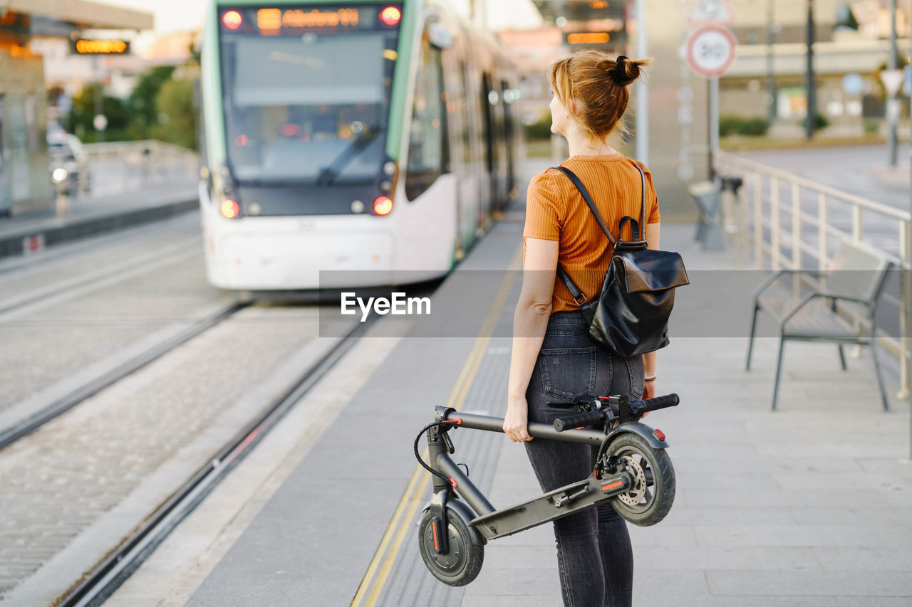 Back view of woman with backpack and folded electric scooter waiting at tram stop