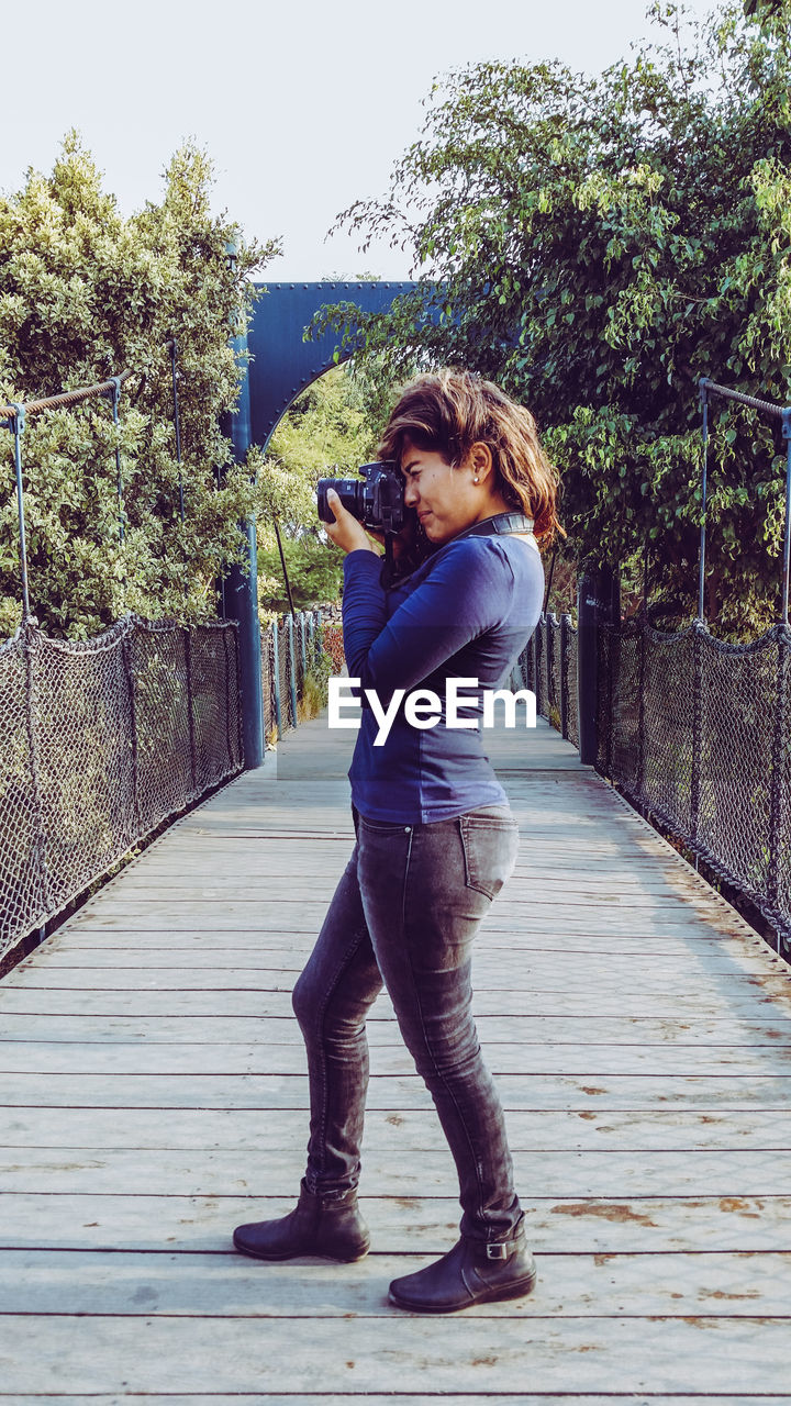 Young woman photographing with camera while standing on footbridge in park