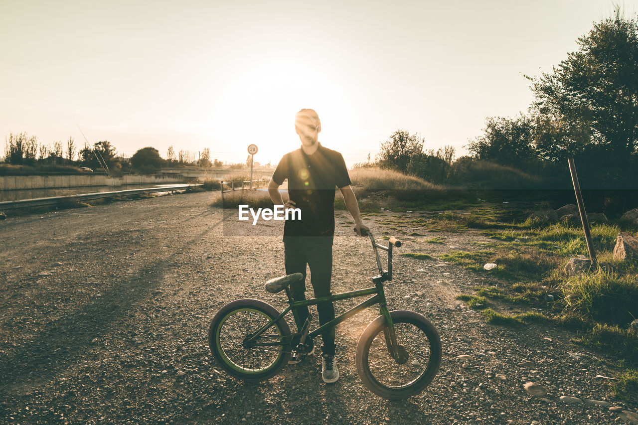 Young man with bicycle standing on field against clear sky during sunset