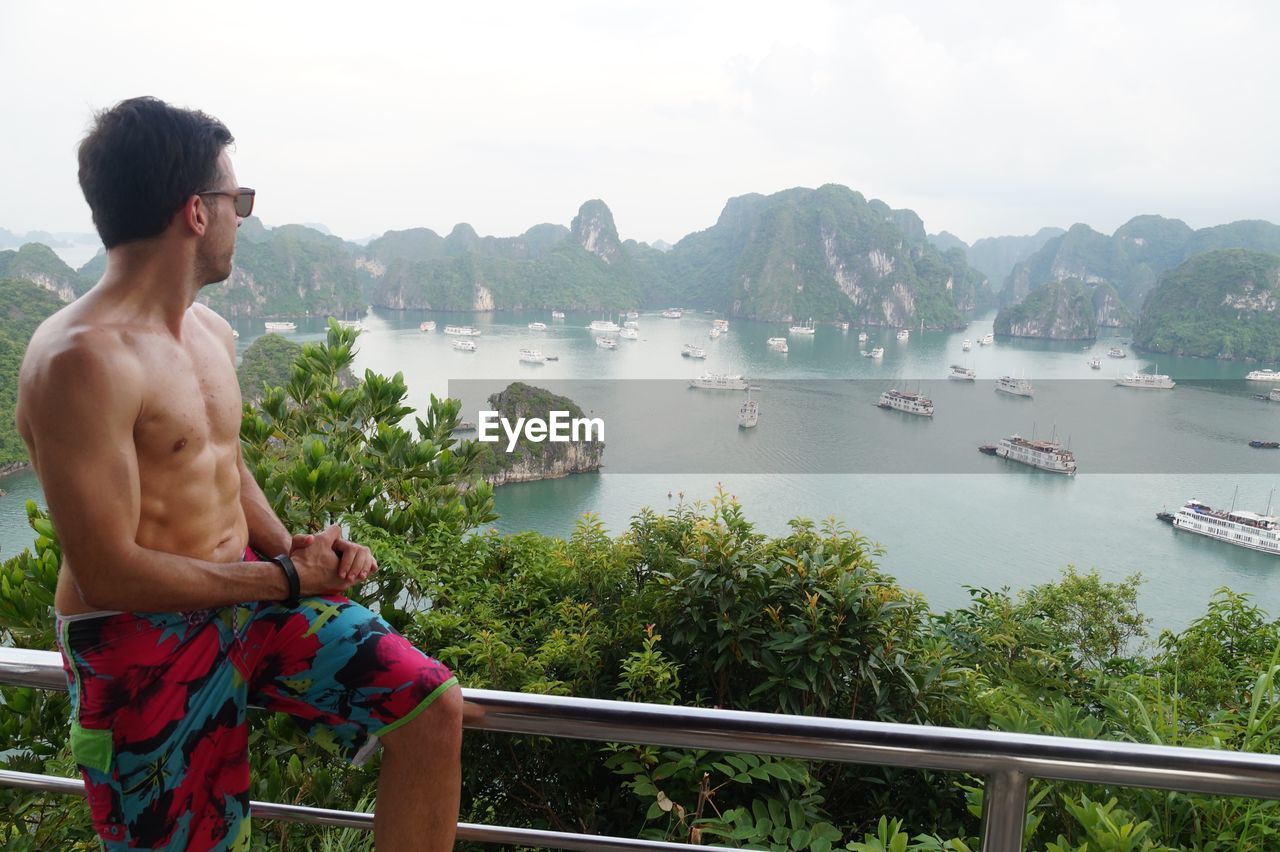 Shirtless man looking at ha long bay while sitting on railing