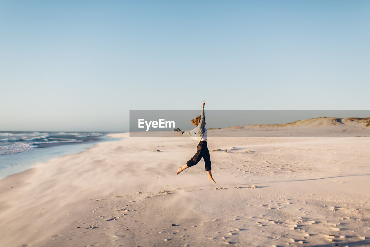 Young woman jumping at beach against clear sky during sunset