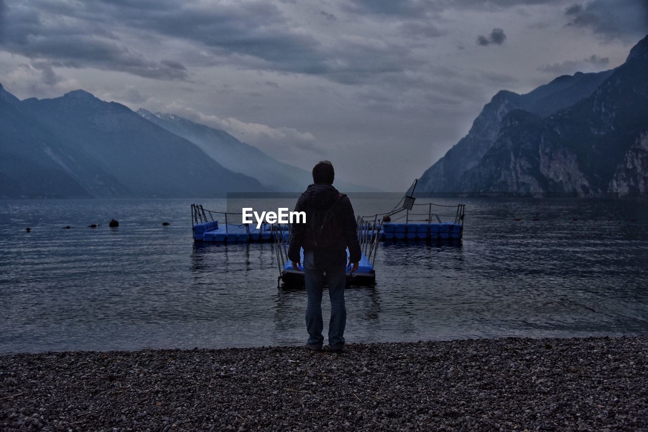 Rear view of man standing on lakeshore against mountains