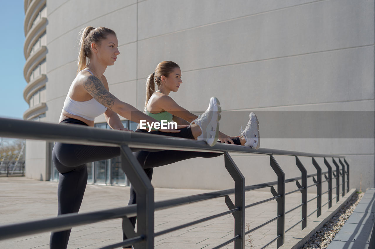 Side view of determined young slim women in sportswear doing stretching exercise for legs while standing near metal railing during outdoor fitness workout in city