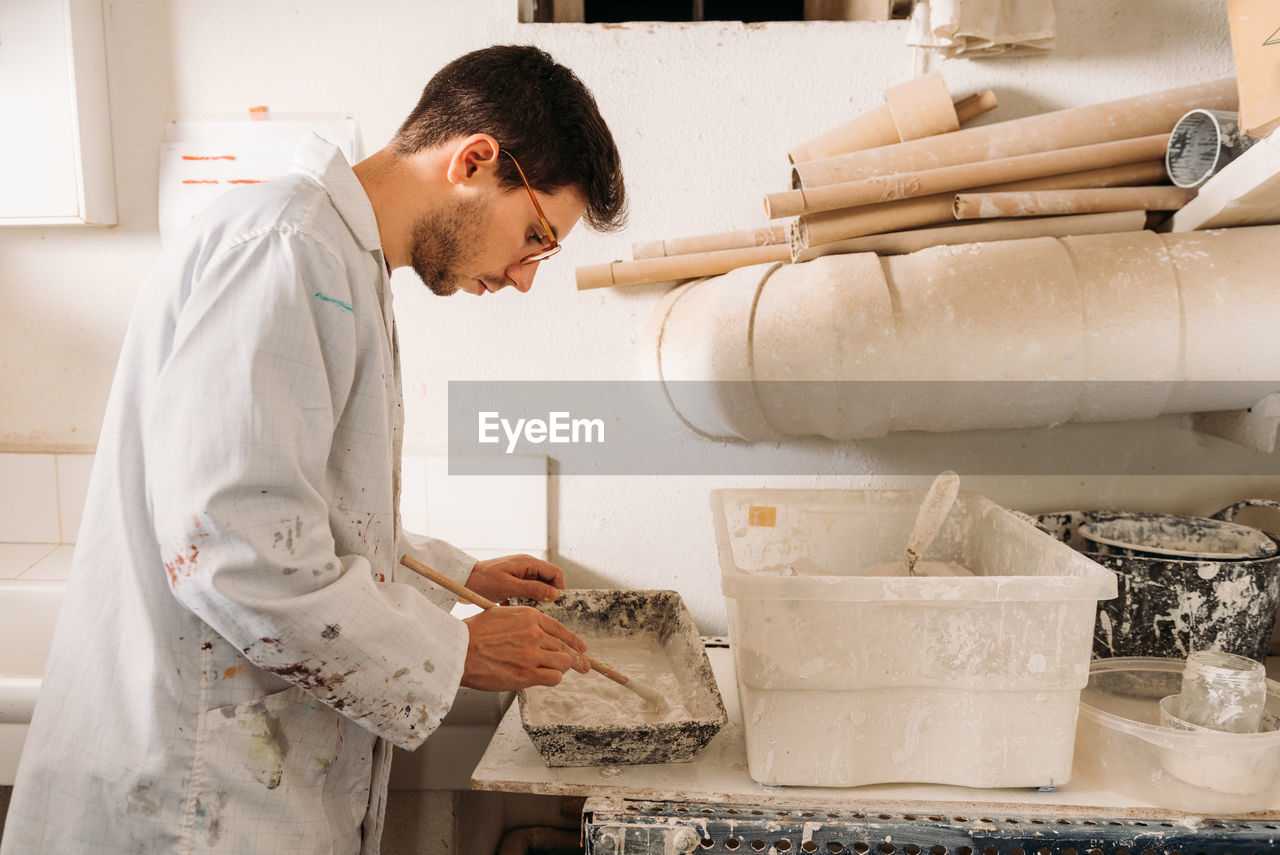 Side view of serious male artisan pouring liquid gypsum in wooden form for shaping material while creating artwork in workshop