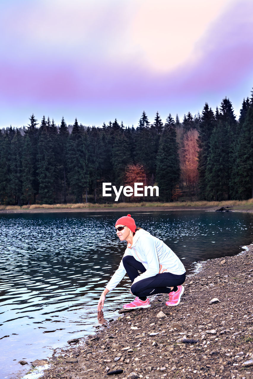 Full length of woman crouching by lake against sky