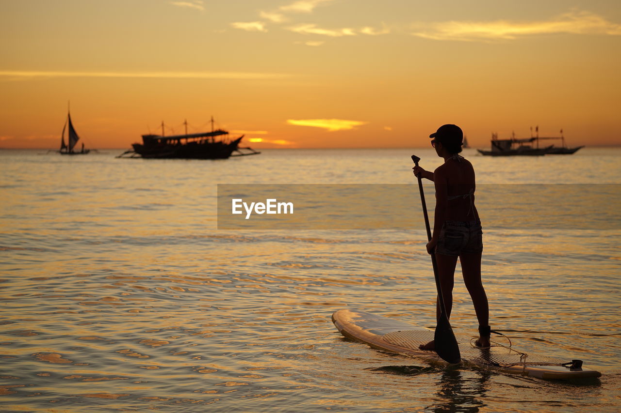 Silhouette woman paddleboarding in sea against orange sky