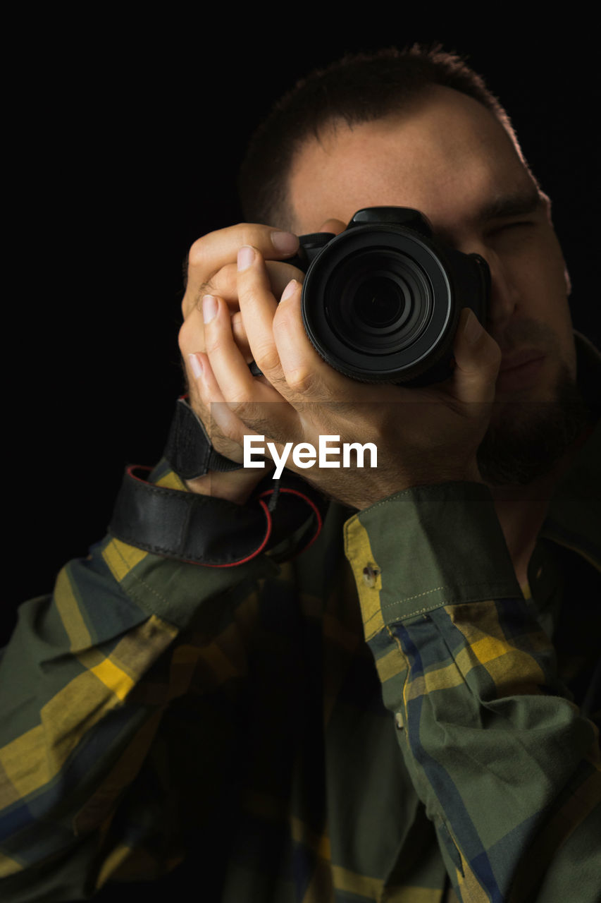 one person, camera, adult, occupation, portrait, technology, black background, activity, studio shot, men, photographer, photographing, holding, costume, person, indoors, clothing, looking through an object, front view, headshot, professional occupation, lens - optical instrument, security, darkness, lens - eye, young adult, obscured face