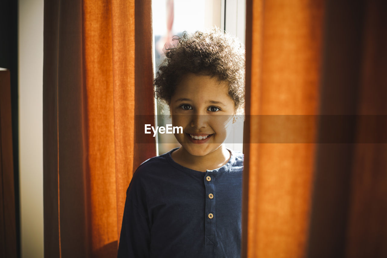 Portrait of smiling boy standing behind curtain at home