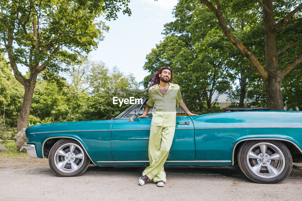 Portrait of young man with legs crossed at ankle standing near vintage car on road in park