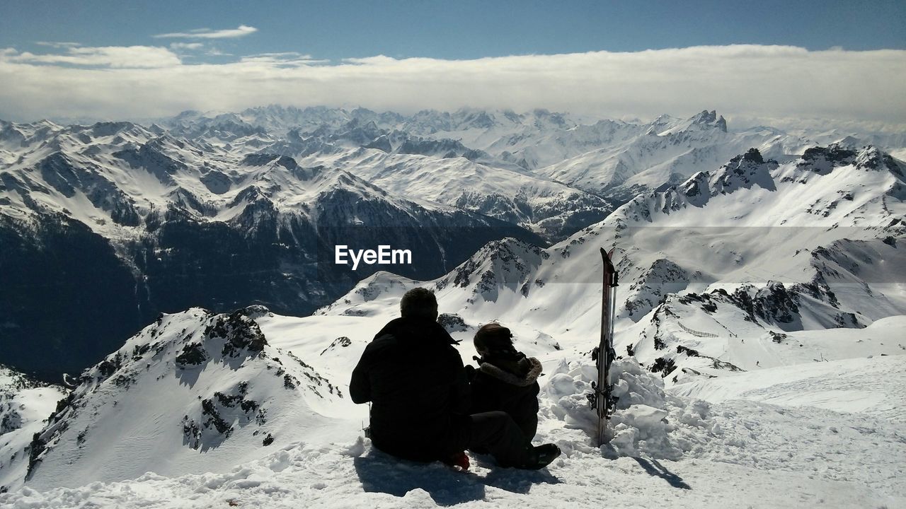 Rear view of friends sitting on snowcapped mountain against sky