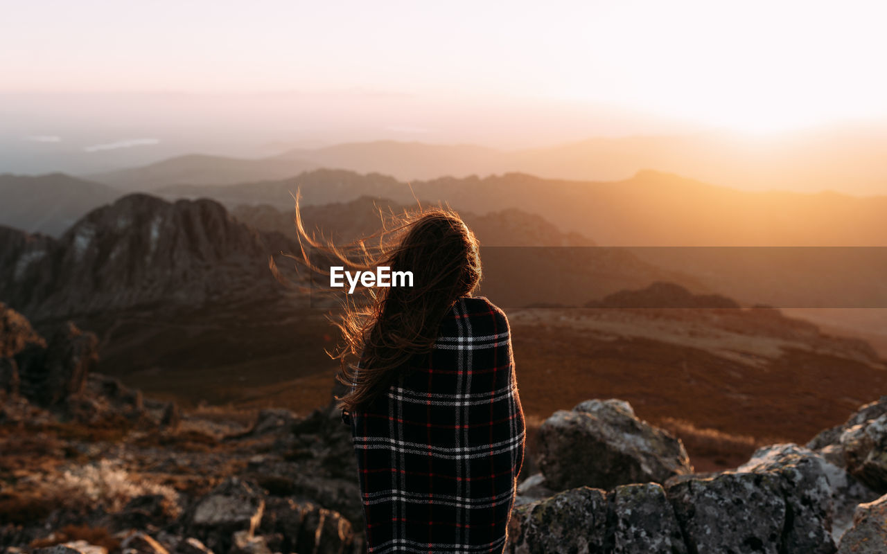 Back view of unrecognizable hiker in blanket standing on stone and observing amazing scenery of highlands valley on sunny day