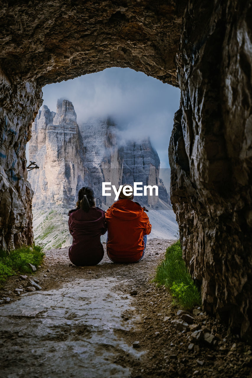 REAR VIEW OF PEOPLE SITTING ON ROCK AGAINST MOUNTAIN