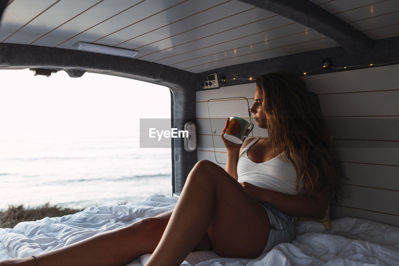 Woman drinking coffee while sitting in camper van at beach