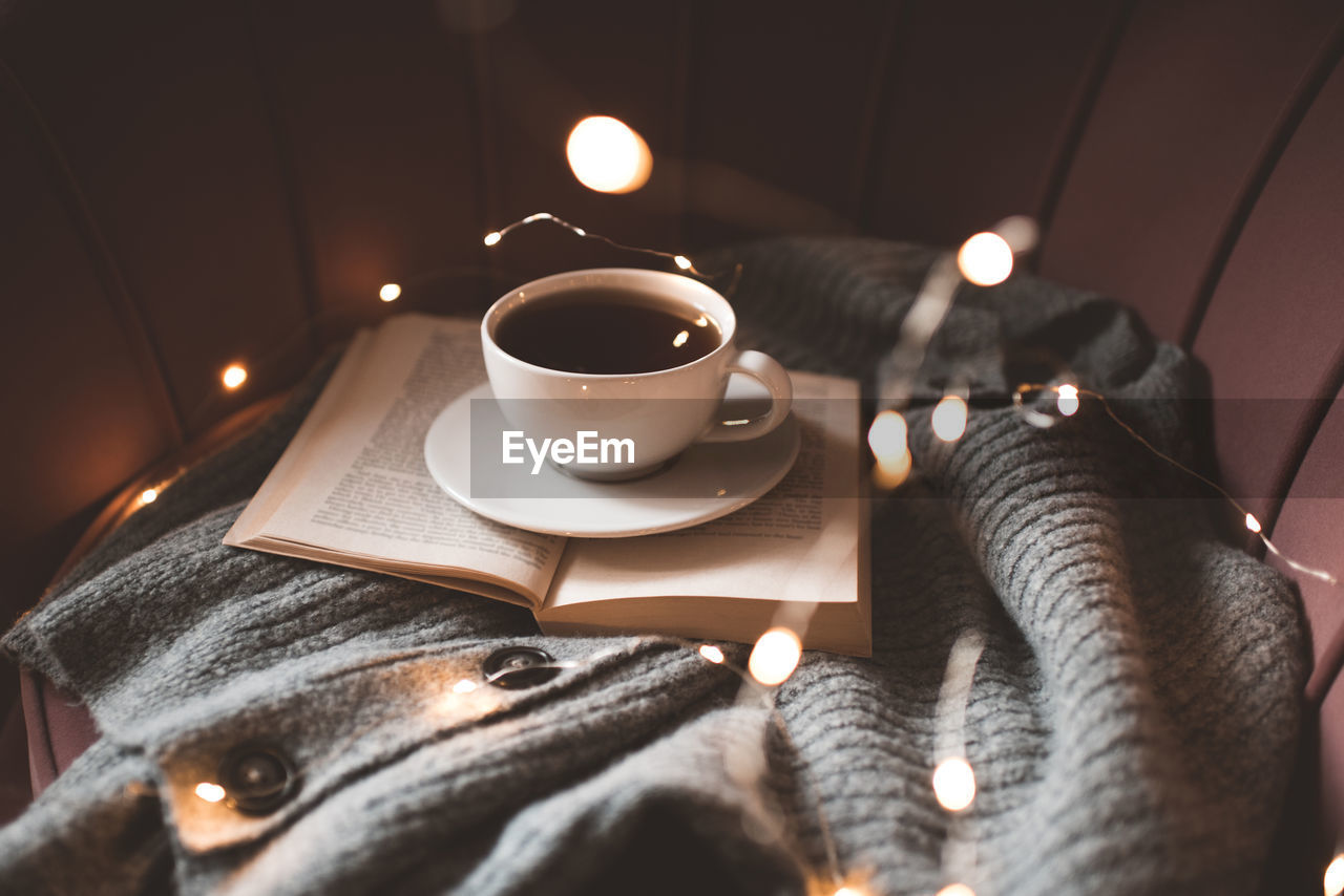 Cup of fresh black tea staying on open paper book with knitted fabric cloth on cozy accent chair 