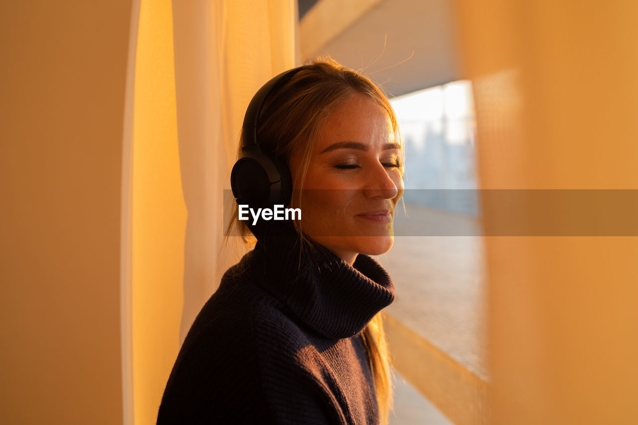 one person, adult, women, listening, portrait, smiling, headphones, indoors, looking, yellow, technology, young adult, happiness, copy space, music, headshot, female, lifestyles, businesswoman, architecture, person, emotion, hairstyle, business, window, wireless technology, brown hair, clothing, standing, side view, arts culture and entertainment, looking away, human face, communication, business finance and industry, cheerful, enjoyment, casual clothing, fashion