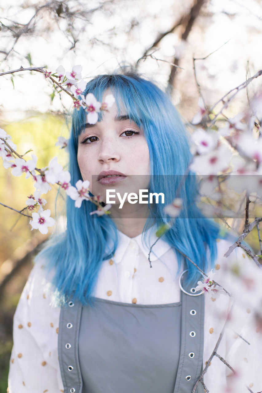 Young stylish female with long blue hair looking at camera wearing trendy overall enjoying blooming tree while standing in spring garden