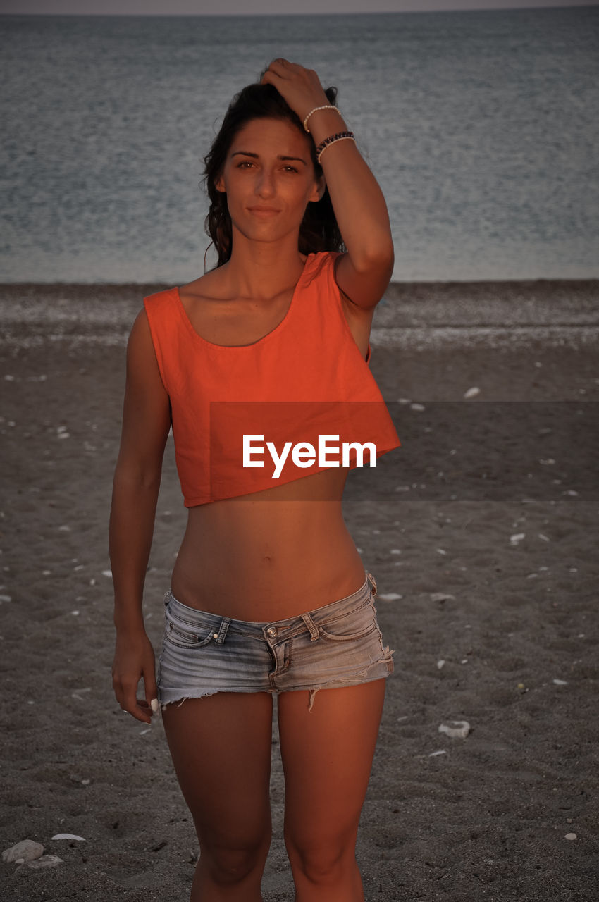 PORTRAIT OF YOUNG WOMAN STANDING AT BEACH