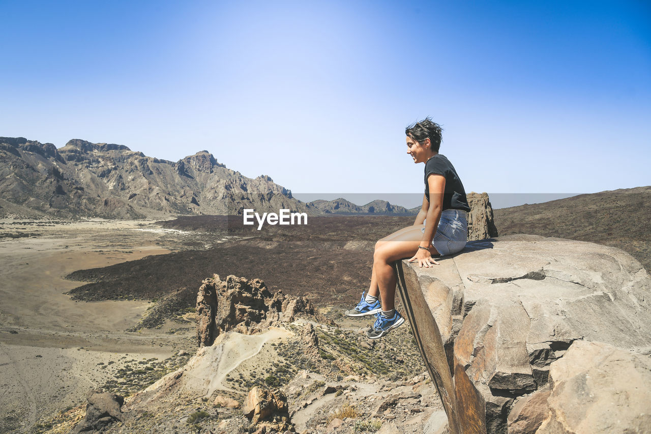 Side view of smiling teenage girl sitting on cliff against blue sky