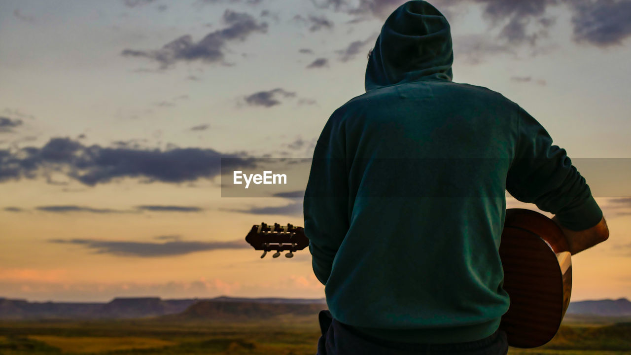 Rear view of man playing guitar against sky during sunset