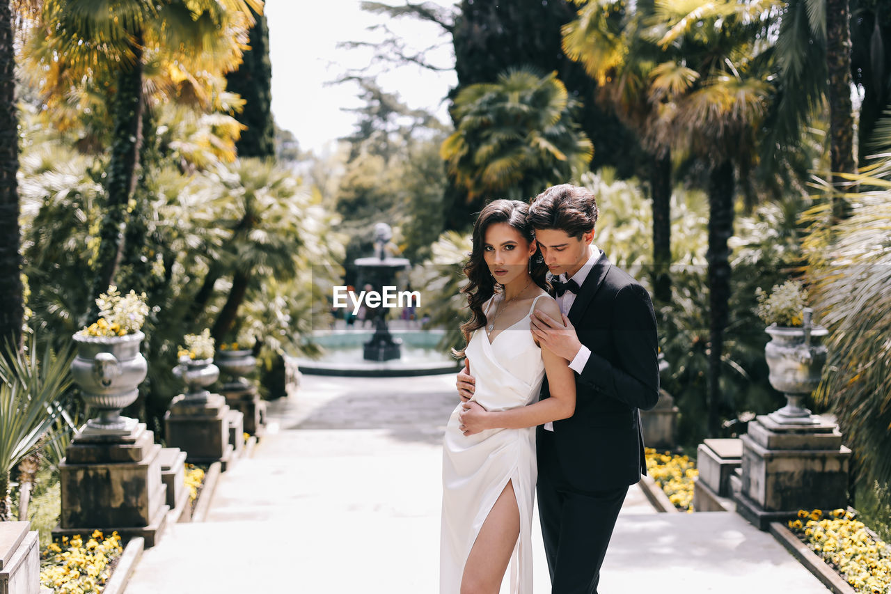 Happy lovers the bride and groom in wedding outfits walk among plants and palm trees in the old park