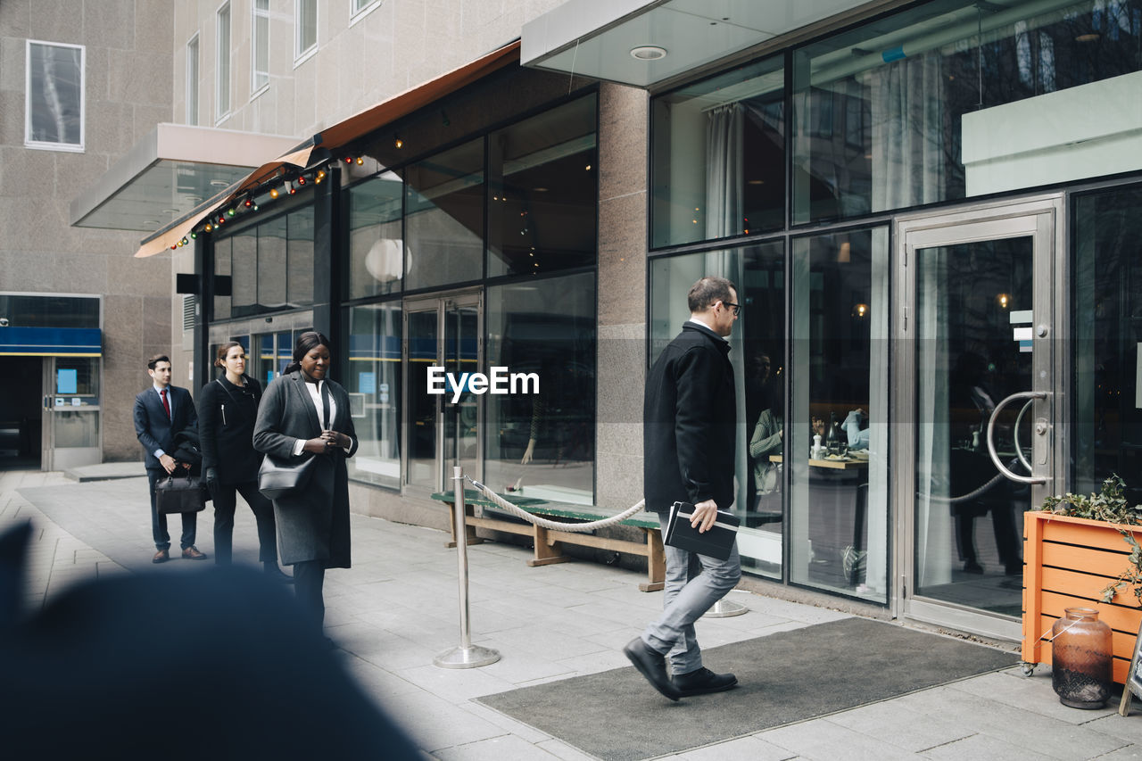 Businessman walking inside store while male and female colleagues standing in line on footpath during covid-19