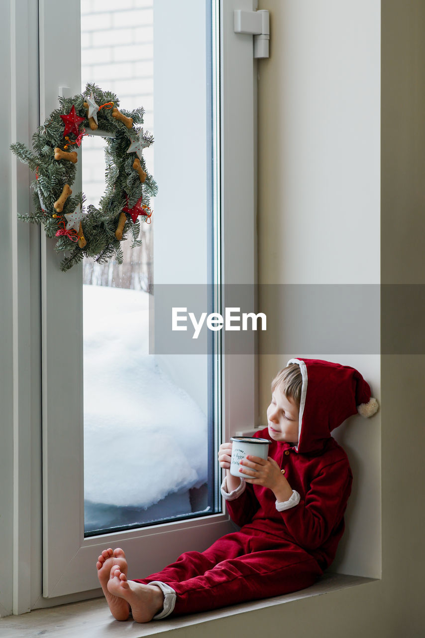 A thoughtful boy in a santa costume sits at the window and makes christmas decorations