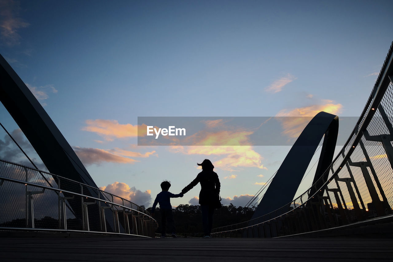Silhouette mother holding hands of son while walking on bridge during sunset