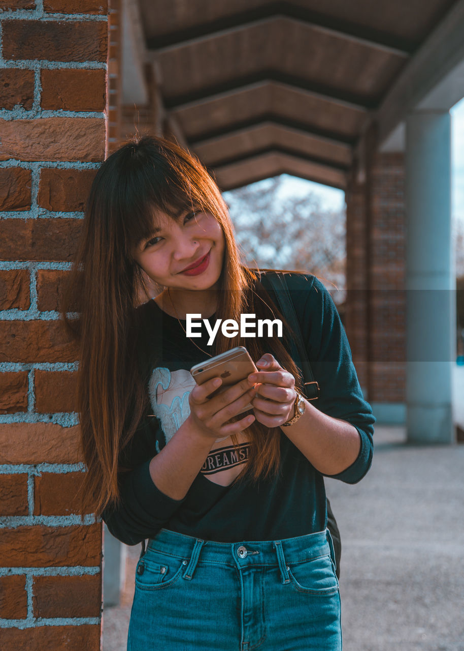 YOUNG WOMAN USING PHONE WHILE STANDING BY BRICK WALL