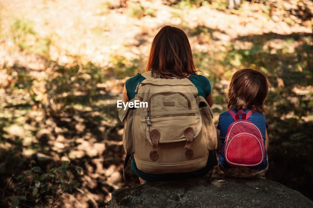 Rear view of mother and daughter with backpacks sitting on rocks in forest