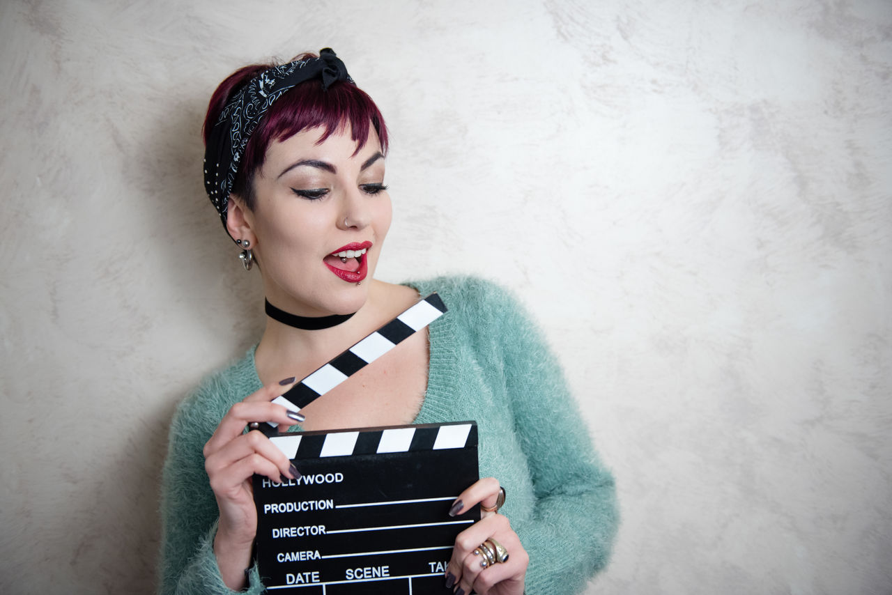 Young woman holding film slate against wall