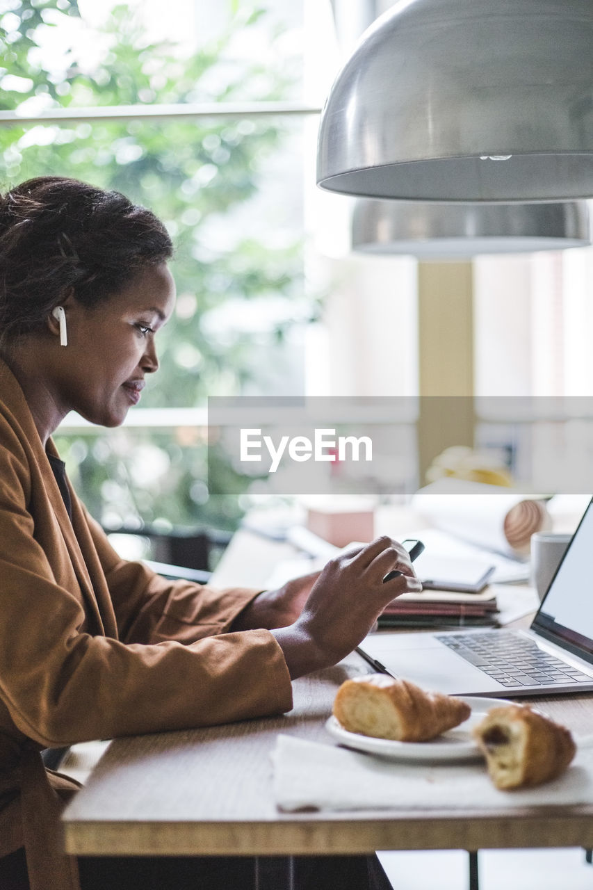 Confident female entrepreneur using phone while sitting by table at home