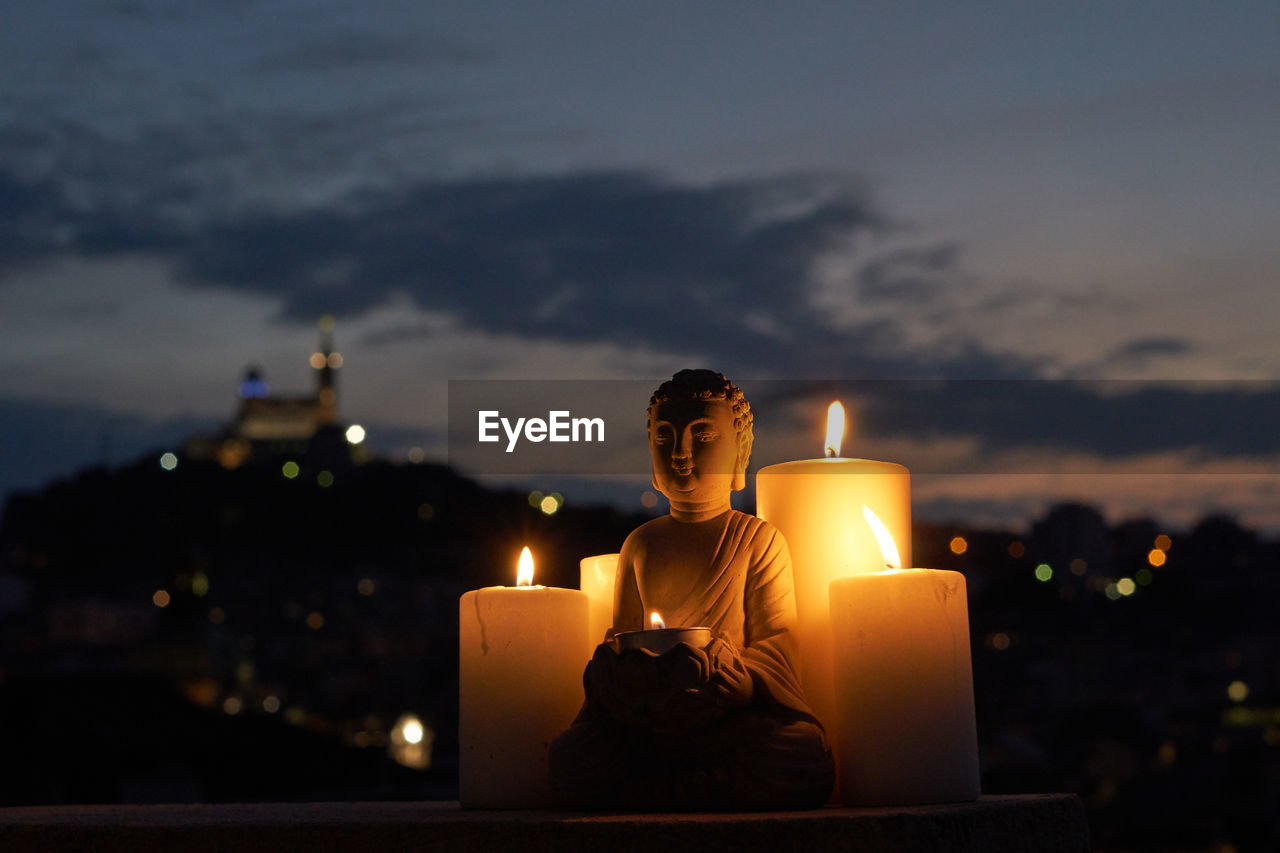 Lit tea light candles and budha against sky at night