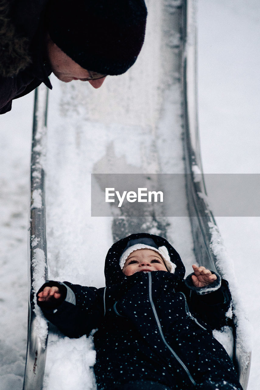 Father looking at baby girl playing on snow covered slide