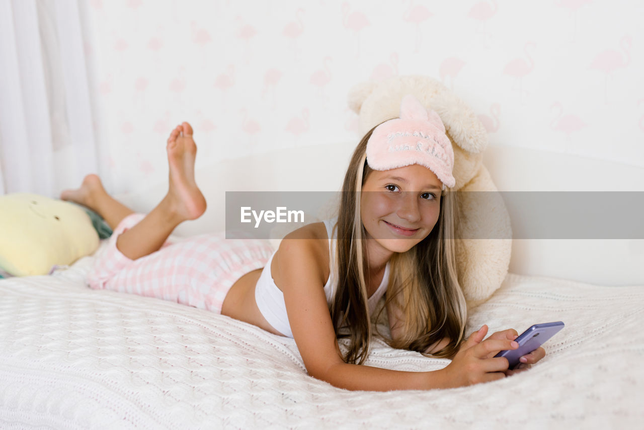 Beautiful teenage girl in pajamas is lying on a cozy bed on her stomach and surfing on the phone