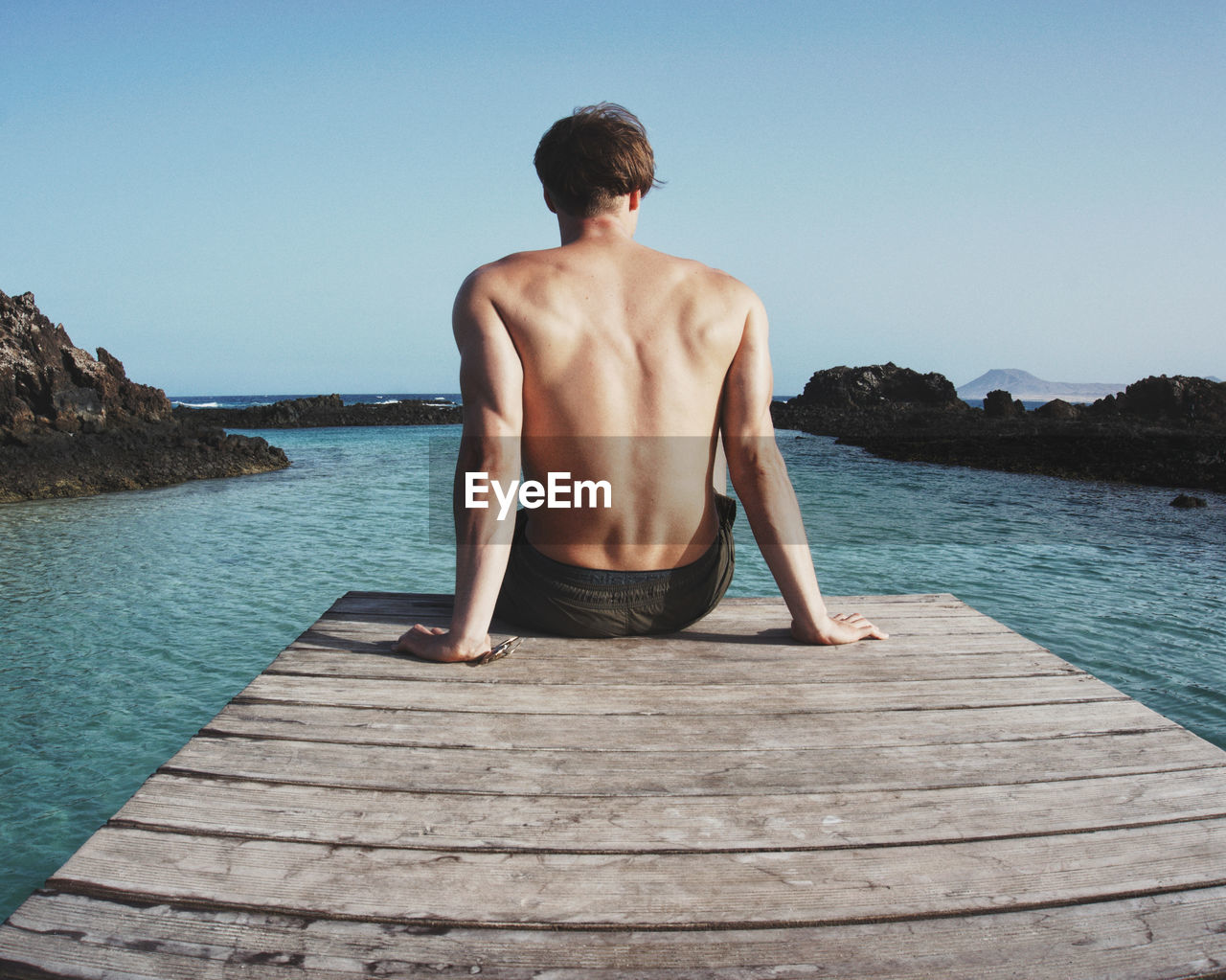 Rear view of shirtless man sitting on pier over sea against clear sky