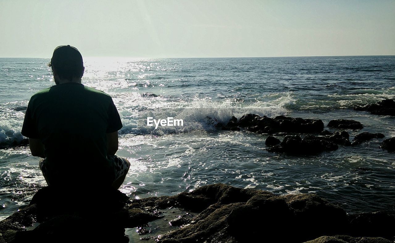 Rear view of man sitting on rock in sea against sky