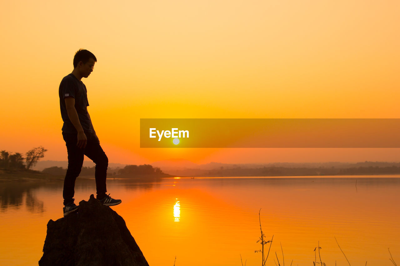 Full length of young man on rock by lake against sky during sunset